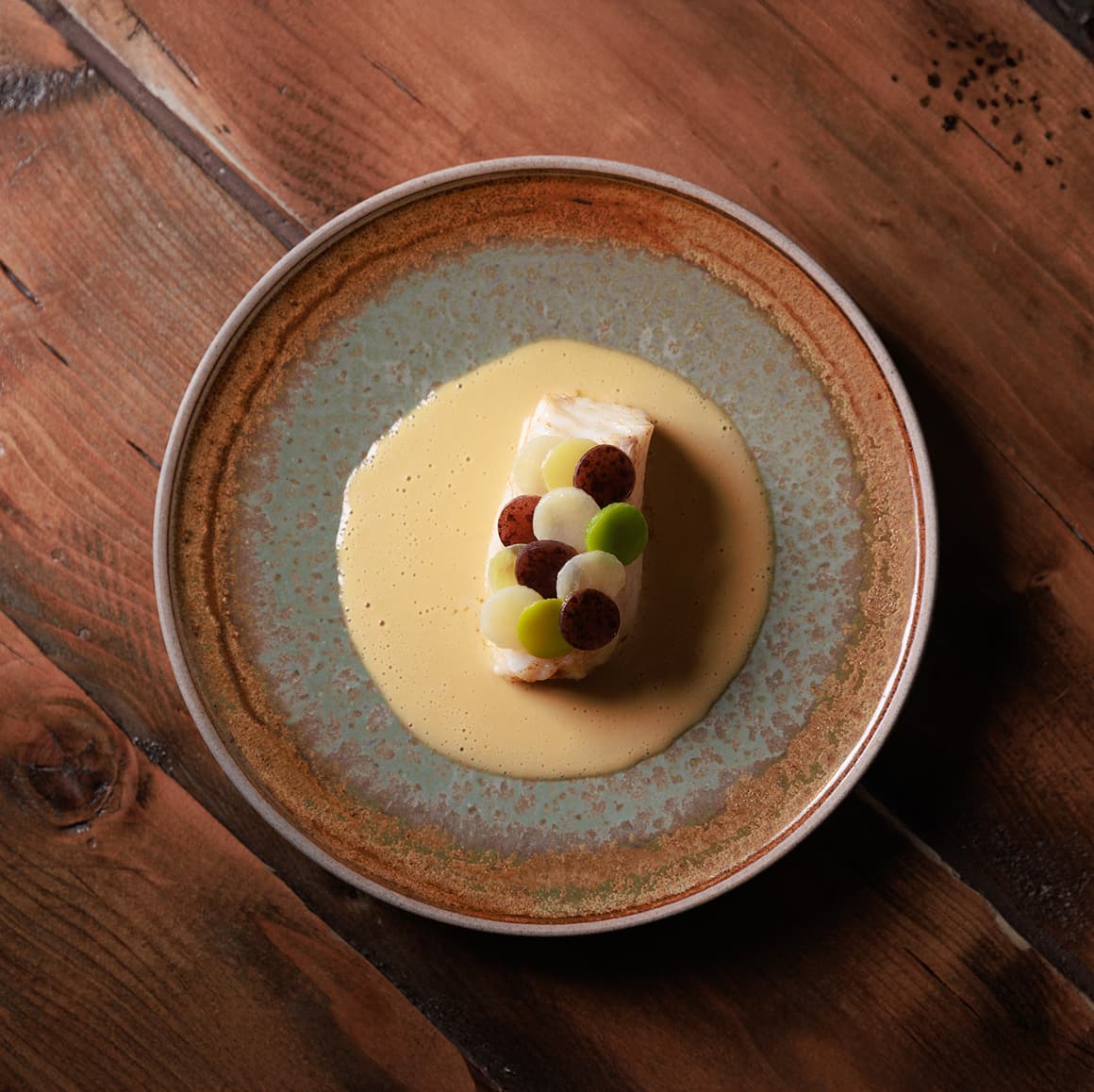 Gourmet dish of layered dessert with cream, garnished with circular toppings, served on a rustic plate with light brown sauce, placed on a wooden table.